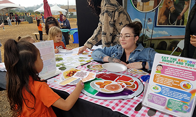A female student kneels to get on a schoolgirl's level as the latter picks food items to put on her MyPlate.
