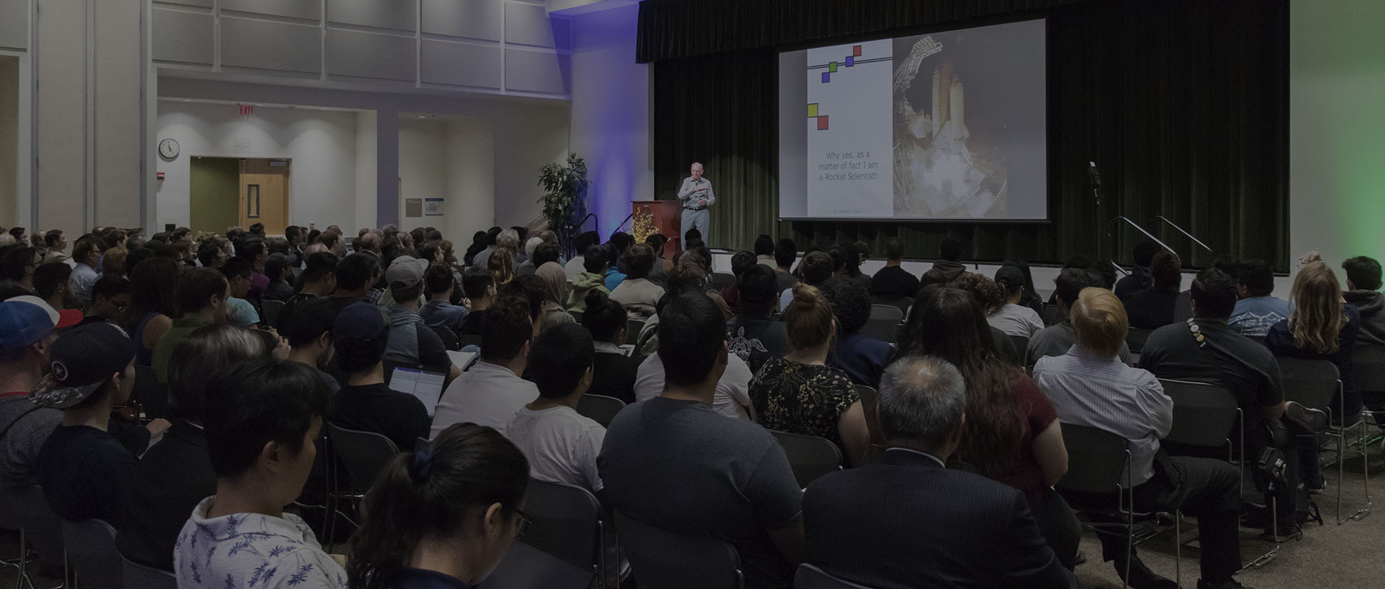 A packed lecture room with a speaker presenting on stage.